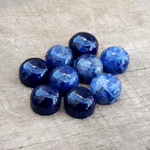 May include: A collection of nine polished, round gemstones in varying shades of blue. The stones have a glossy finish, reflecting light, and are arranged on a weathered wooden surface. The gemstones appear to be natural stones.