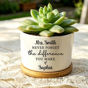 Puede incluir: Una maceta de cerámica blanca con una planta suculenta. La maceta tiene el texto "Mrs. Smith never forget the difference you make. Sophia" en escritura negra. Se encuentra sobre una base redonda de madera.