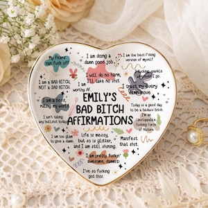Könnte beinhalten: Eine herzförmige weiße Keramikschale mit goldenem Rand, die den schwarzen Schriftzug EMILY'S BAD BITCH AFFIRMATIONS trägt, umgeben von verschiedenen bunten, handgezeichneten Kritzeleien, kleinen Formen und zusätzlichen motivierenden Sprüchen.