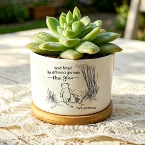 Puede incluir: Una maceta de cerámica blanca con una planta suculenta, con una ilustración de Winnie the Pooh y el texto "Never forget the difference you make Mrs. Glow". La maceta está sobre un platillo de madera.