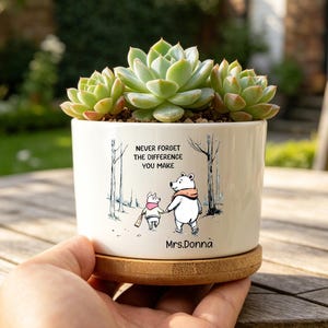 Puede incluir: Una maceta de cerámica blanca con una planta suculenta, con una ilustración de Winnie the Pooh y el texto "NEVER FORGET THE DIFFERENCE YOU MAKE". La maceta está sobre un platillo de madera.