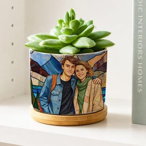 May include: A cylindrical ceramic planter featuring a colorful stained-glass style illustration of a person in a denim jacket and another in a beige coat, topped with a green succulent and resting on a circular bamboo wood base.