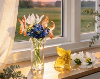 Könnte beinhalten: Eine klare Glasvase mit gewelltem Rand enthält einen Strauß aus handgefertigten Glasblumen in Blau, Weiß und Orange mit grünen Stielen. Zwei weitere Glasblumen, eine gelbe und eine weiße, liegen auf der hölzernen Oberfläche.