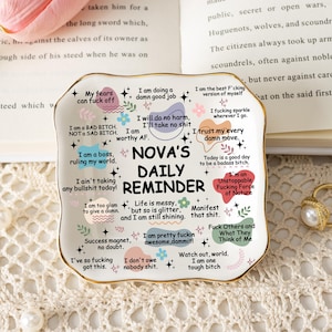 Könnte beinhalten: Ein weißer Keramik-Tageserinnerungsteller mit goldenem Rand, der verschiedene positive Affirmationen und den Text "NOVA'S DAILY REMINDER" enthält. Der Teller ist mit Blumen- und Sternmotiven verziert.
