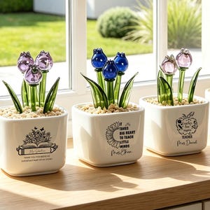 May include: Four square white ceramic pots containing glass tulip flowers in purple, blue, and amber colors. Each pot features black printed text and illustrations, including floral designs and teacher-themed quotes, filled with small light-colored pebbles.
