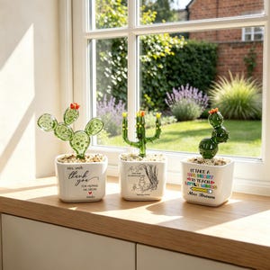 May include: Three glass cactus figurines in white square ceramic pots filled with small stones. The first pot says Mrs. Smith thank you for helping me grow. The second features a Winnie the Pooh illustration. The third says It take a big heart to teach little minds Mrs. Brown.