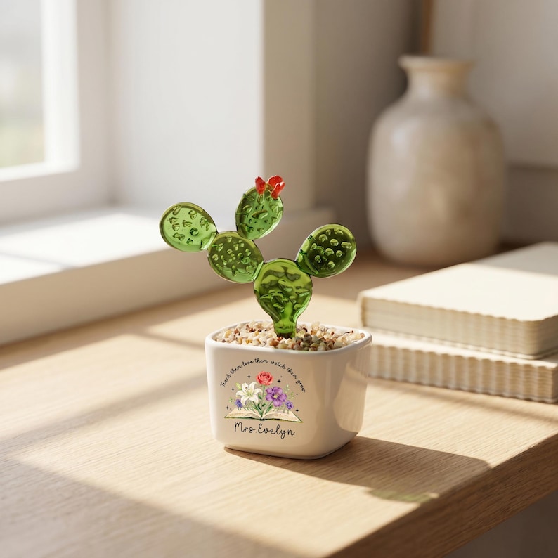 May include: A green glass cactus with a small red flower on top, planted in a white square ceramic pot filled with small pebbles. The pot features a floral graphic, an open book illustration, and the text "Teach them love them watch them grow" and "Mrs Evelyn".