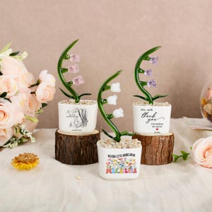 May include: Three glass lily of the valley ornaments in white square pots filled with small stones. Each pot features unique text and graphics, including Winnie the Pooh, thank you messages, and floral designs, all displayed on individual wood slices.