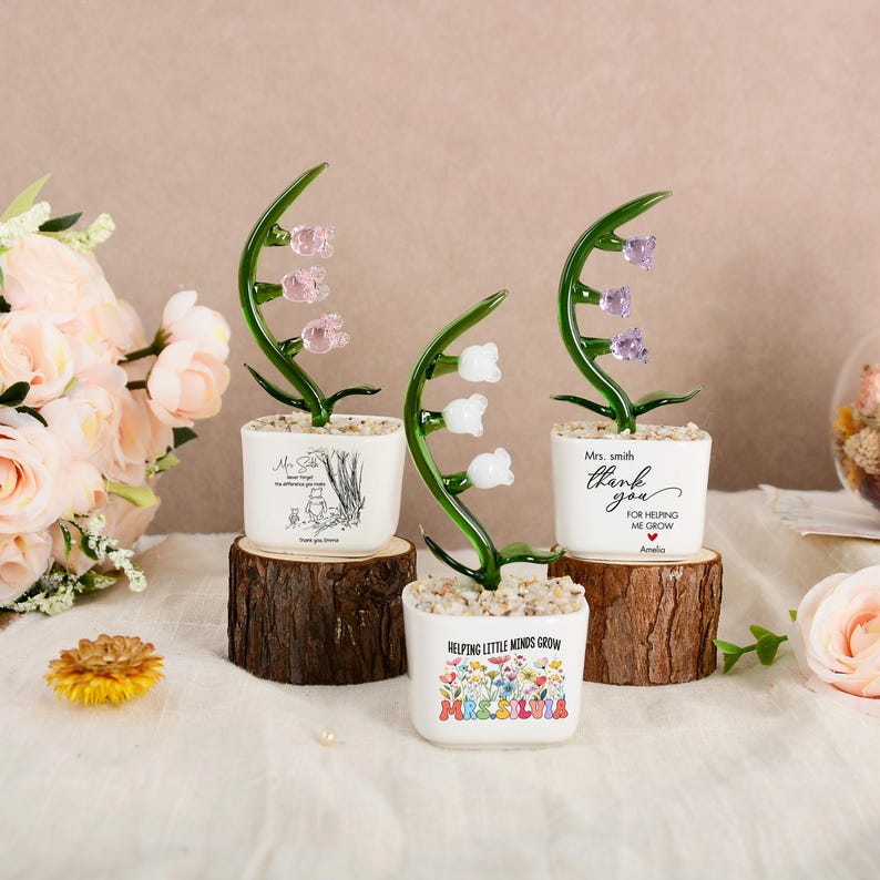 May include: Three glass lily of the valley ornaments in white square pots filled with small stones. Each pot features unique text and graphics, including Winnie the Pooh, thank you messages, and floral designs, all displayed on individual wood slices.