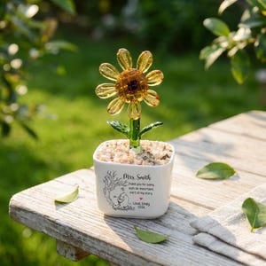 May include: A glass sunflower with yellow petals and a green stem planted in a white square ceramic pot filled with small stones. The pot features a black printed illustration of a bear and text reading Mrs. Smith Thank you for being such an important part of my story. Love, Emily 2026.