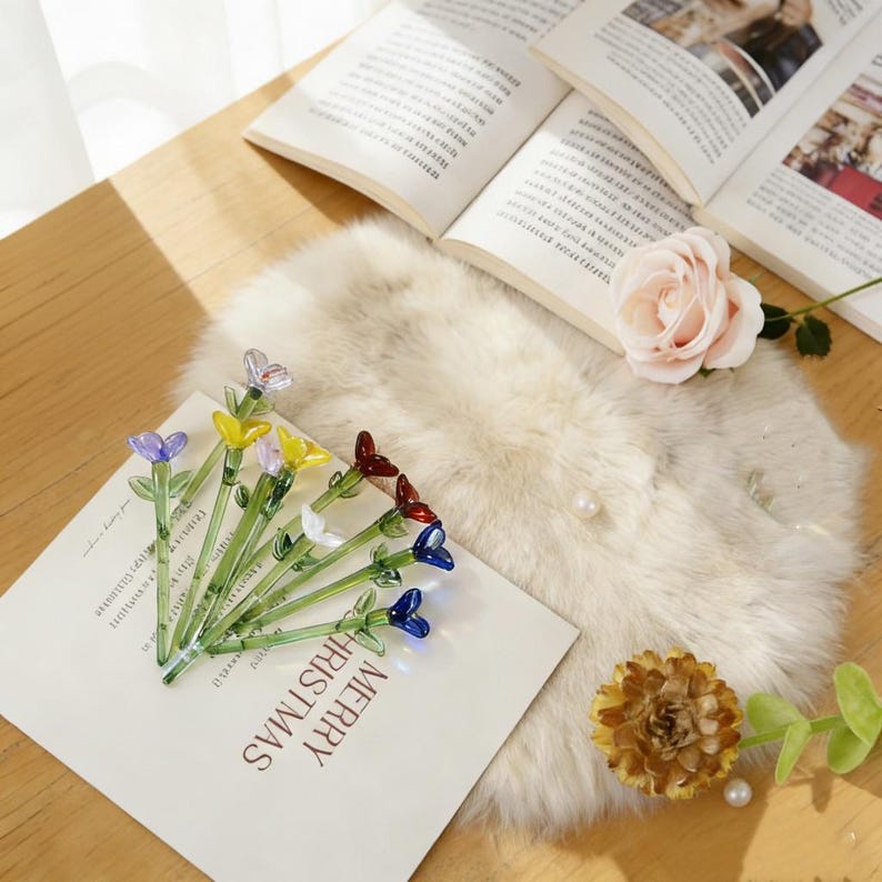 Pu&ograve; includere: Una collezione di fermagli per capelli a forma di fiori in vetro colorato, con steli verdi e fiori blu, rossi, gialli e bianchi. I fermagli sono disposti su una carta bianca con la scritta "MERRY CHRISTMAS".