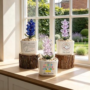 May include: Three white ceramic planters on wooden log bases, each containing a glass flower sculpture. The planters feature printed text and graphics, including 'Mrs. Smith', 'Love, Emily 2021', 'It take a big heart to teach little minds', and 'Mrs. Brown'.