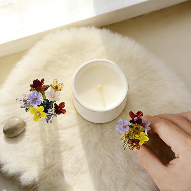 Pu&ograve; includere: Una candela bianca in un contenitore rotondo &egrave; circondata da accessori per capelli a forma di fiori di vetro colorati. Un accessorio &egrave; tenuto in mano. I fiori sono rossi, gialli, viola e trasparenti.