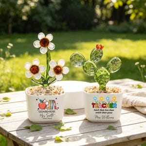 May include: Two white square ceramic planters containing glass plant sculptures. The left planter features a red apple, books, and the text TEACH LOVE INSPIRE Mrs. Smith From Paula. The right planter has a cactus and the text TEACHER teach them love them watch them grow Mrs. Williams.