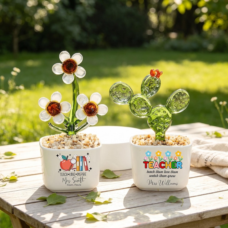 May include: Two white square ceramic planters containing glass plant sculptures. The left planter features a red apple, books, and the text TEACH LOVE INSPIRE Mrs. Smith From Paula. The right planter has a cactus and the text TEACHER teach them love them watch them grow Mrs. Williams.