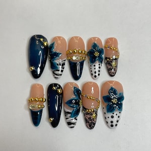 Könnte beinhalten: Ein Set aus zehn künstlichen Nägeln mit verschiedenen Designs. Die Nägel weisen marineblaue, nudefarbene und weiße Basen auf, verziert mit goldenen Akzenten, floralen Verzierungen und komplizierten Mustern. Einige Nägel haben ein glänzendes Finish.
