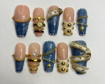 May include: A set of ten artificial nails with a variety of designs. The nails feature a combination of light pink and blue colors, adorned with gold chains, beads, and star accents. Some nails have a French tip design.