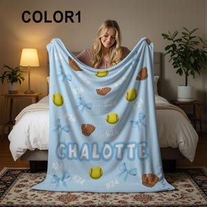 May include: A light blue softball blanket with a personalized name, softball graphics, and the number 24. The blanket features yellow softballs, brown gloves, and light blue bows. The name "CHALOTTE" is displayed in large, white letters.