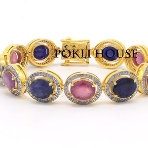 Op de afbeelding: Gouden armband met ovale edelstenen in blauw en roze, geaccentueerd met kleine, heldere stenen. De armband heeft een sluiting en wordt gepresenteerd tegen een witte achtergrond. Het is een sieraad.