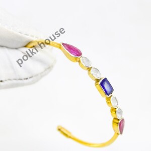 Könnte beinhalten: Ein goldfarbenes Armreif-Armband mit einer Reihe farbiger Edelsteine. Das Armband hat eine Kombination aus roten, weißen und blauen Steinen, die in einzelnen goldenen Fassungen gefasst sind. Das Armband wird von einer behandschuhten Hand gehalten.