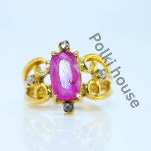 May include: A gold ring featuring a central rectangular faceted pink gemstone held by prongs, flanked by smaller dark stones on a decorative openwork band. The text Polki house is printed diagonally in grey next to the ring on a white background.