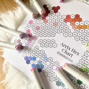 May include: A collection of Arrtx markers and a color chart. The markers are white with colored tips, scattered around a paper with a hexagonal color chart. The chart is labeled "Arrtx Hex Chart" and includes color codes.