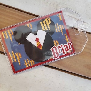 May include: A clear plastic luggage tag with a red border. The tag features a blue background with a Mickey Mouse head wearing a white shirt and a red and gold tie. The background is covered with the letters "HP" in a repeating pattern. The name "Briar" is printed in white letters on the tag.