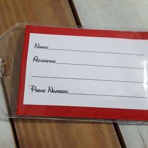 May include: A red luggage tag with a clear plastic cover. The tag has white lines for writing "Name", "Address", and "Phone Number".