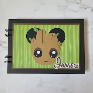 May include: A black notebook with a green and brown striped cover. The cover features a cartoon character with a brown face, large eyes, and green leaves on its head. The name "James" is written in brown cursive on the cover.