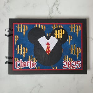 May include: Black scrapbook with a blue and gold Harry Potter themed cover. The cover features a Mickey Mouse head with a black suit and red and gold tie. The text "Charlie 2025" is on the cover.