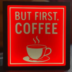 Può includere: Un cartello quadrato rosso con testo bianco che recita "BUT FIRST, COFFEE". Sotto il testo c'è una tazza di caffè bianca con vapore che sale. Il cartello ha un bordo nero.