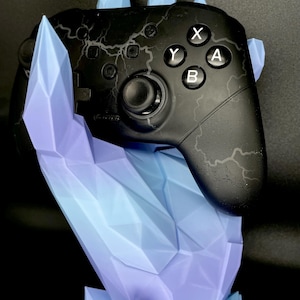 Könnte beinhalten: Ein schwarzer Videospiel-Controller mit Blitzmuster, der auf einem blau-violetten kristallförmigen Ständer ruht. Der Controller hat weiße X-, Y-, A- und B-Tasten.