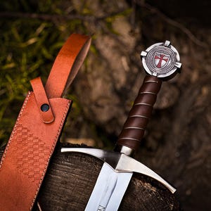 May include: A medieval-style sword with a brown leather-wrapped handle and a silver blade. The hilt features a circular emblem with a red cross. A matching brown leather sheath is visible.