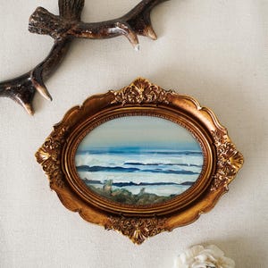 May include: An oval framed painting of a seascape with blue and white waves. The ornate gold frame is surrounded by a brown antler and a white flower-shaped candle.