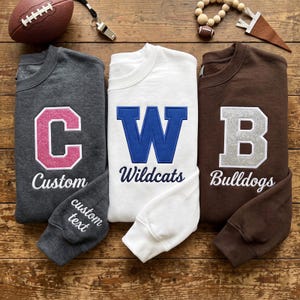 Peut inclure: Trois sweat-shirts à col rond en gris anthracite, blanc et marron. Le sweat-shirt gris présente une lettre C à paillettes roses et le texte Custom. Le blanc a une lettre W bleue et Wildcats. Le marron a une lettre B à paillettes argentées et Bulldogs.