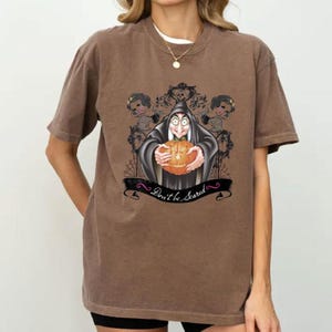May include: Brown t-shirt featuring a Halloween-themed graphic. The design includes a cartoon witch holding a carved pumpkin, surrounded by decorative elements and the text "Don't be scared".