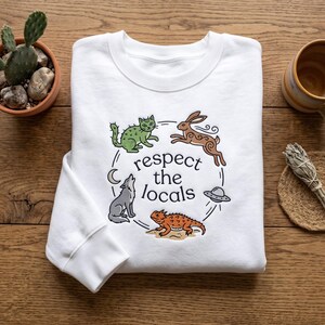 May include: A white long-sleeve sweatshirt featuring a graphic print with the text "respect the locals" surrounded by illustrations of a green cactus cat, a brown rabbit, a gray wolf, a horned lizard, a crescent moon, and a flying saucer.