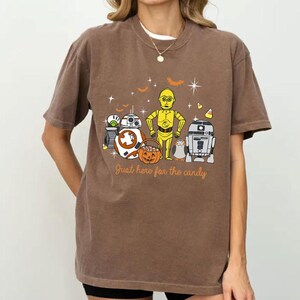 Peut inclure: T-shirt marron avec un motif d'Halloween sur le thème de Star Wars. Le design comprend divers personnages, une citrouille, des chauves-souris et le texte "Just here for the candy".
