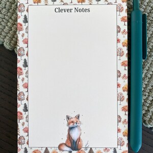 May include: A notepad with the text "Clever Notes" and a watercolor illustration of a fox. The notepad has a border of autumn trees in shades of orange, red, and brown. A teal pen rests beside the notepad.