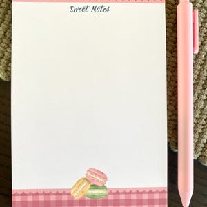 May include: A pink pen rests beside a notepad with a pink gingham border and the words "Sweet Notes." The notepad features a stack of watercolor macarons in pink, yellow, and green. The background is a textured, neutral color.