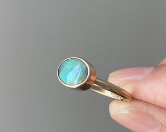 May include: A gold ring featuring a round, iridescent opal stone. The opal displays shades of blue, green, and white. The ring's band is thin and appears to be made of gold. The ring is held up by a hand.