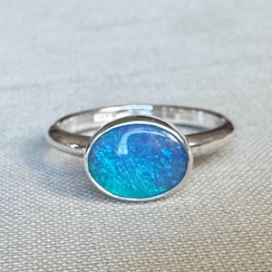 May include: A silver ring with an oval-shaped, iridescent blue opal stone. The opal displays shades of blue and green. The ring is set on a light-colored surface.
