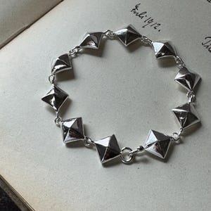 Op de afbeelding: Een zilverkleurige kettingarmband bestaande uit tien individuele piramidevormige schakels verbonden door kleine ronde ringetjes. Elke schakel heeft een gepolijst, reflecterend oppervlak met vier driehoekige facetten.