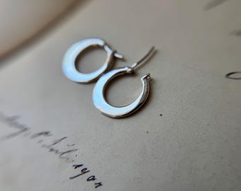 May include: A pair of small, polished silver hoop earrings with a smooth, rounded surface. Each earring features a thin post and a secure hinge mechanism, showcasing a reflective metallic finish that catches the light on its curved, circular frame.