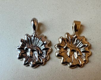 May include: Two pendants with a floral design. One pendant is silver-colored, and the other is gold-colored. Each pendant has a loop at the top for a chain. The pendants are on a light beige surface.
