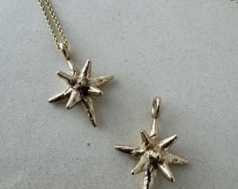 May include: A gold-toned necklace featuring a star-shaped pendant with a textured, uneven surface, accompanied by a separate matching star-shaped pendant charm. Both pieces exhibit a multi-pointed design with a central raised point.