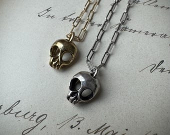 May include: Two skull pendant necklaces. One is gold-toned, the other silver-toned, each suspended from a chain. The pendants are detailed with eye sockets, a nose cavity, and teeth. The necklaces rest on a vintage paper background.