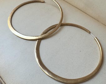 May include: A pair of large, circular hoop earrings with a brushed gold finish. The earrings feature a thin, metallic wire post for closure and have a slightly hammered, textured surface that catches the light along the curved edges.