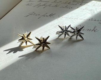 May include: Two pairs of star-shaped stud earrings, one pair in a polished gold tone and one pair in a polished silver tone. Each earring features a central point with multiple radiating spikes, creating a dimensional, multi-pointed star design.
