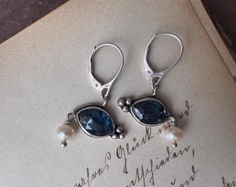 May include: A pair of silver earrings with dark blue, oval-shaped gemstones and small, dangling white pearls. The earrings have a lever-back closure and are set against a vintage paper background.